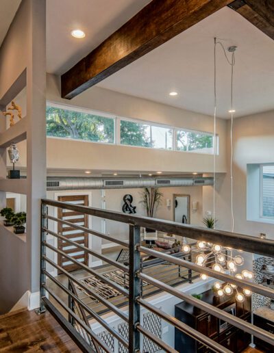 A modern home interior featuring a high ceiling with wooden beams, a balcony with metal railings overlooking a dining area, and large windows for natural lighting.
