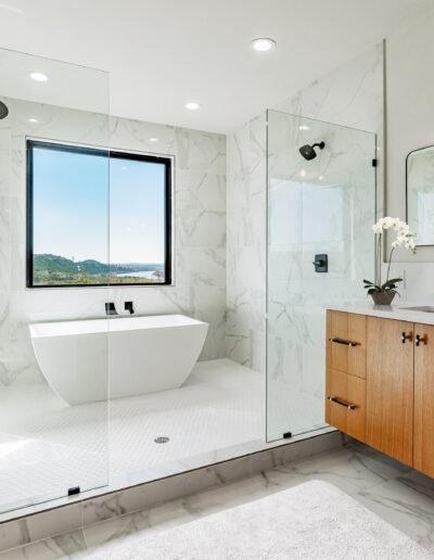Modern bathroom with marble tiles featuring a freestanding tub, glass-enclosed shower, and wooden vanity under a large mirror with a scenic view through the window.