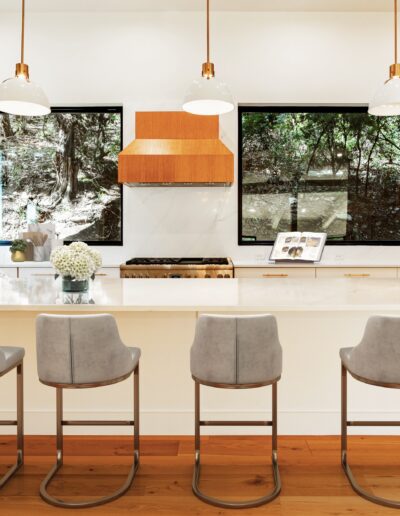 Modern kitchen interior with white countertops, three bar stools, pendant lights, and a view of the outdoors through a large window.