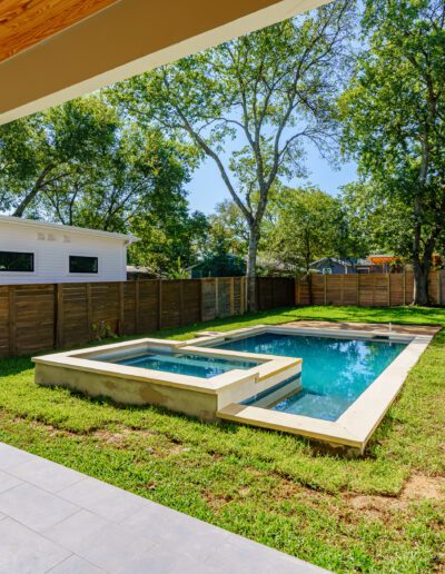 Backyard with a small rectangular pool and hot tub, surrounded by grass and enclosed by a wooden fence, with a patio area and a modern single-story building in the background.