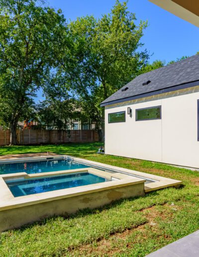 Modern backyard with a small pool and a well-kept lawn next to a white house with a slanted roof.
