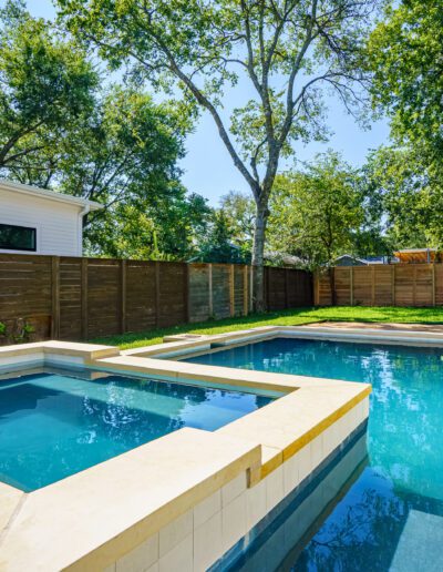 Backyard with a modern rectangular swimming pool and an adjacent hot tub beside a wooden fence on a sunny day.