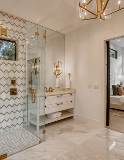 A modern bathroom with a glass shower enclosure, white and gold tile walls, and a view into an adjoining bedroom.