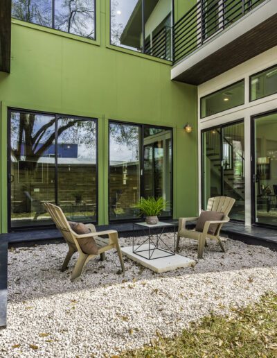 Modern backyard patio with chairs and a small table on a gravel surface, adjacent to a green contemporary house.