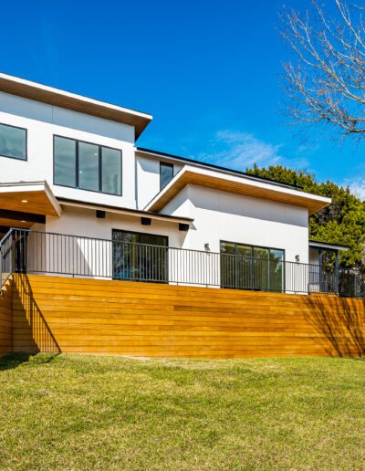 Modern two-story house with a wooden deck and a fenced backyard on a sunny day.