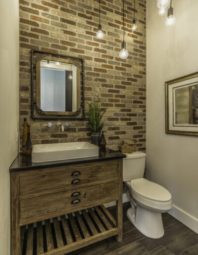 A modern bathroom featuring an exposed brick wall, wooden vanity, hanging light bulbs, and a framed mirror.