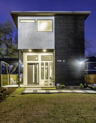 Modern two-story home illuminated at twilight with a dark brick façade, landscaped front yard, and wooden fence.