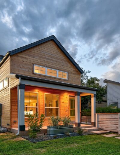 Modern two-story home with illuminated interior at dusk, featuring wooden siding and a fenced-in yard.