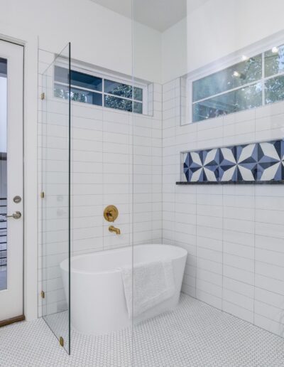 A bright, modern bathroom with white subway tiles, a freestanding bathtub, and a glass shower, featuring a door leading outside and decorative plants.