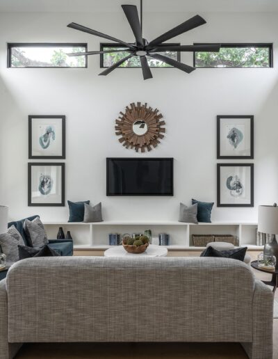 Modern living room with high ceilings, large windows, a ceiling fan, a grey sofa, wall art, a wall-mounted TV, and a symmetrical arrangement of decor. White walls and wood floors enhance the airy feel.
