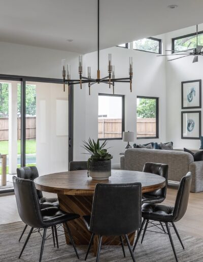Modern open-concept living area with a dining table, sectional sofa, and large windows showing a backyard. Decor includes framed artwork, a wall-mounted TV, and a chandelier.