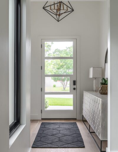 A modern entryway with a geometric light fixture, a black rug on light wood flooring, a narrow console table with a lamp, and a glass front door offering a view of greenery outside.