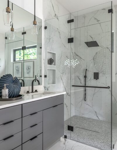 Modern bathroom with a large glass-enclosed shower, white freestanding bathtub, double sink vanity with grey cabinets, and a marble countertop. Decor includes plants and framed artwork.