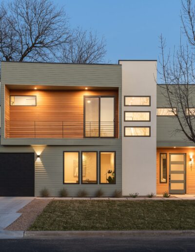 Modern two-story house with clean lines, large windows, and a mix of wood, white, and gray exterior surfaces. The front yard has minimal landscaping and a driveway leading to a garage. Evening lighting.