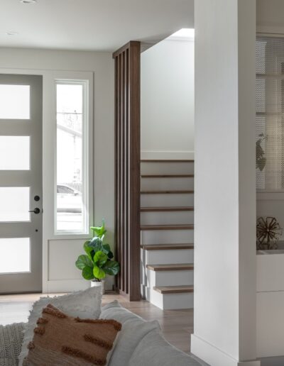 A modern and bright entryway with a gray front door, a plant, a staircase, and a view into a living room with white cabinetry and a sofa.