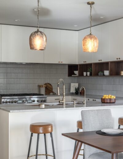 Modern kitchen with white cabinetry, gray tile backsplash, marble countertops, a gas stove, and a double sink. Three pendant lights hang over the island, and a dining table with chairs is in the foreground.