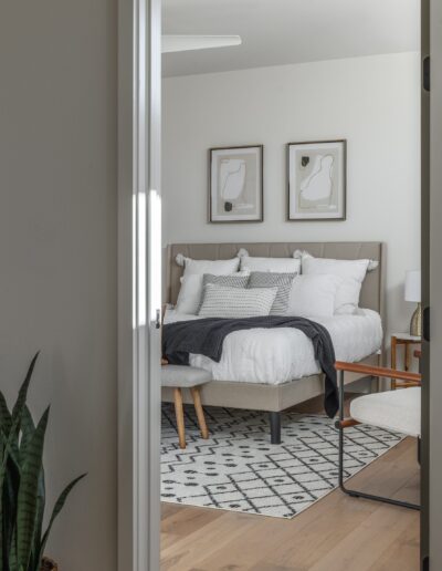 A modern bedroom with a white bed, gray blanket, two framed abstract artworks, a patterned rug, a plant, and minimalistic furniture, viewed through an open door.