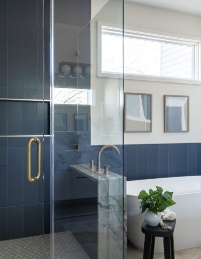 A modern bathroom featuring blue-tiled walls, a glass-enclosed shower, a freestanding bathtub, a vanity with two sinks, and framed art above the tub. A small plant sits on a stool next to the tub.